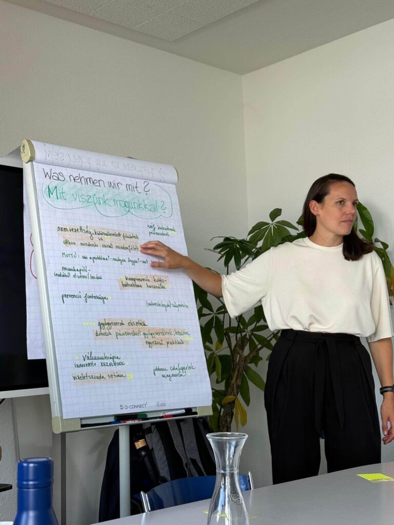 Egy nő rámutat a flipchart táblára, amelyen a Mit viszünk magunkkal kérdésre adott válaszok vannak felírva a keddi műhelymunka során
